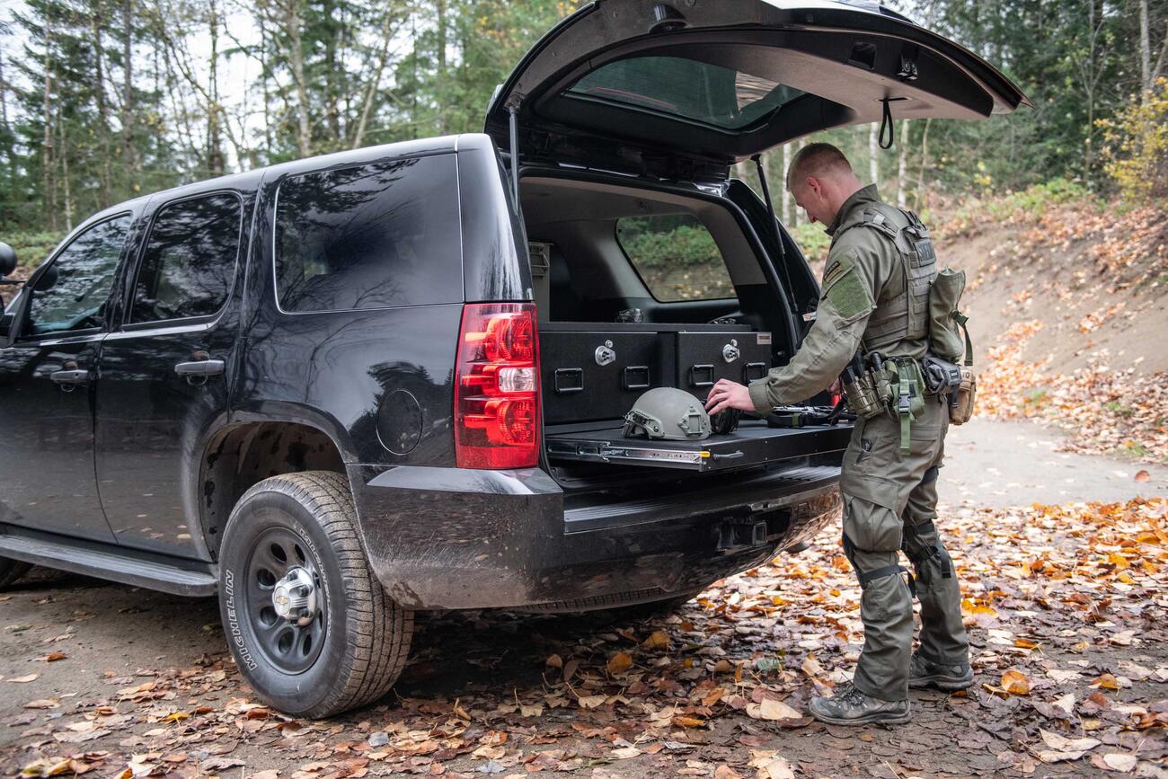 A man in tactical clothing working on a gun that is on top of a heavy duty pull out table in the back of a black Chevy Tahoe.