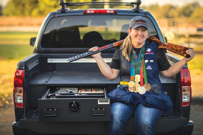 Kim Rhode sitting on the tailgate of her Nissan Titan while holding her Olympic gold medals and competition rifle.