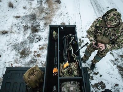 A man in camouflage checking his phone next to a TruckVault with a compound bow inside of it.