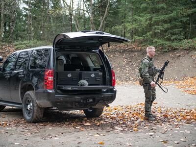 A man in tactical clothing holding a gun walking behind a black Chevy Tahoe complete with a TruckVault and a heavy duty pull out table.