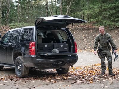 A man in tactical clothing holding a gun walking behind a black Chevy Tahoe complete with a TruckVault and a heavy duty pull out table.