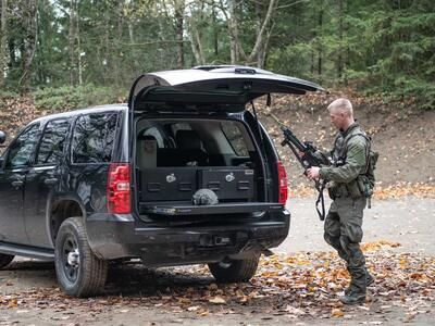 A man in tactical clothing holding a gun walking behind a black Chevy Tahoe complete with a TruckVault and heavy duty pull out table.