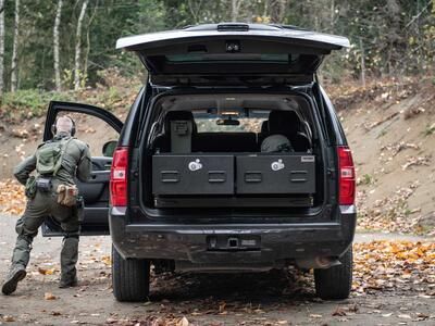 A man in tactical clothing taking cover behind a black Chevy Tahoe with a TruckVault inside of it.