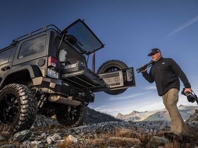 A man holding a camera and tripod near a black jeep with a TruckVualt in the mountains 