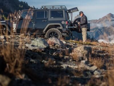 A man changing out a camera lense behind a black 2013 Jeep Wrangler with a TruckVault and a heavy duty pull out table in the mountains.