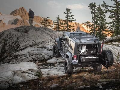 A gray Jeep Wrangler with a custom TruckVault parked on rocks in a mountain range with a man overlooking a valley.