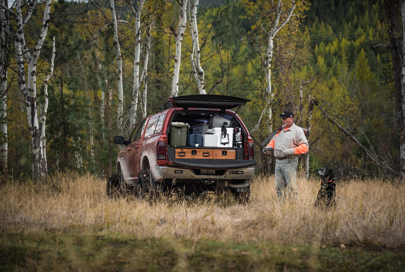 bird hunter and black dog stand in tall grass with covered Dodge Ram pickup and TruckVault secure storage