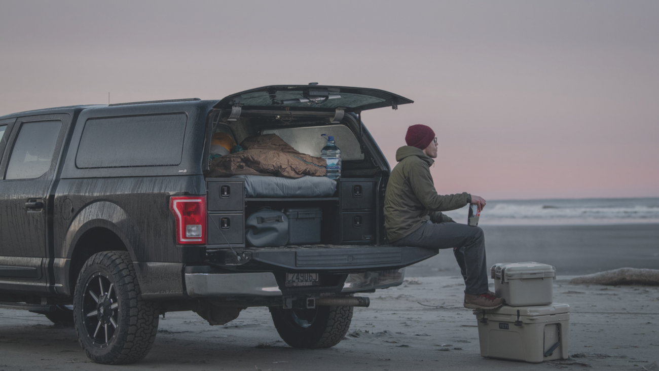 Black Ford F-150 with bed cap and tailgate open showing a TruckVault Base Camp 4. Man sitting on tailgate with feet resting on coolers watching the sun set at a beach