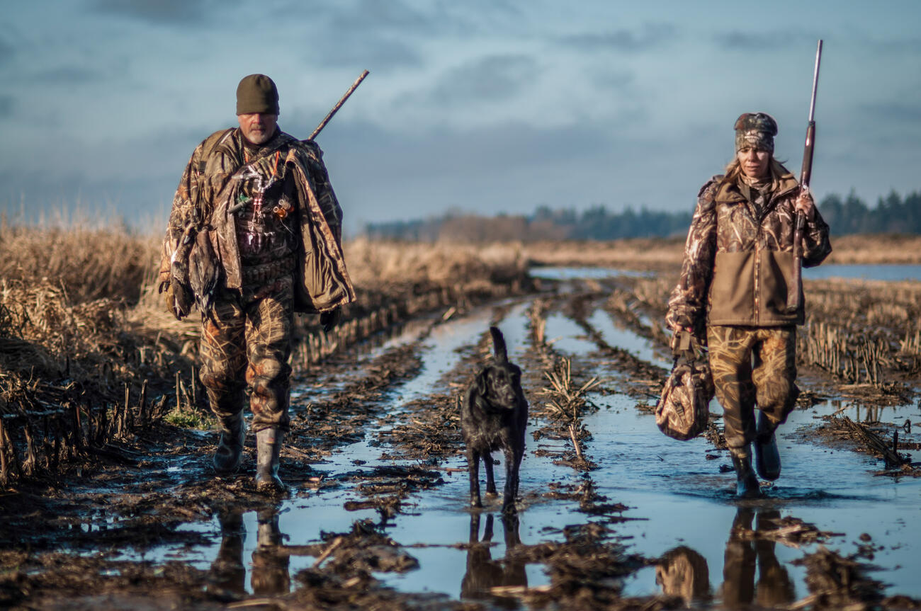 two hunters with black lab in middle walking towards camera through a marsh