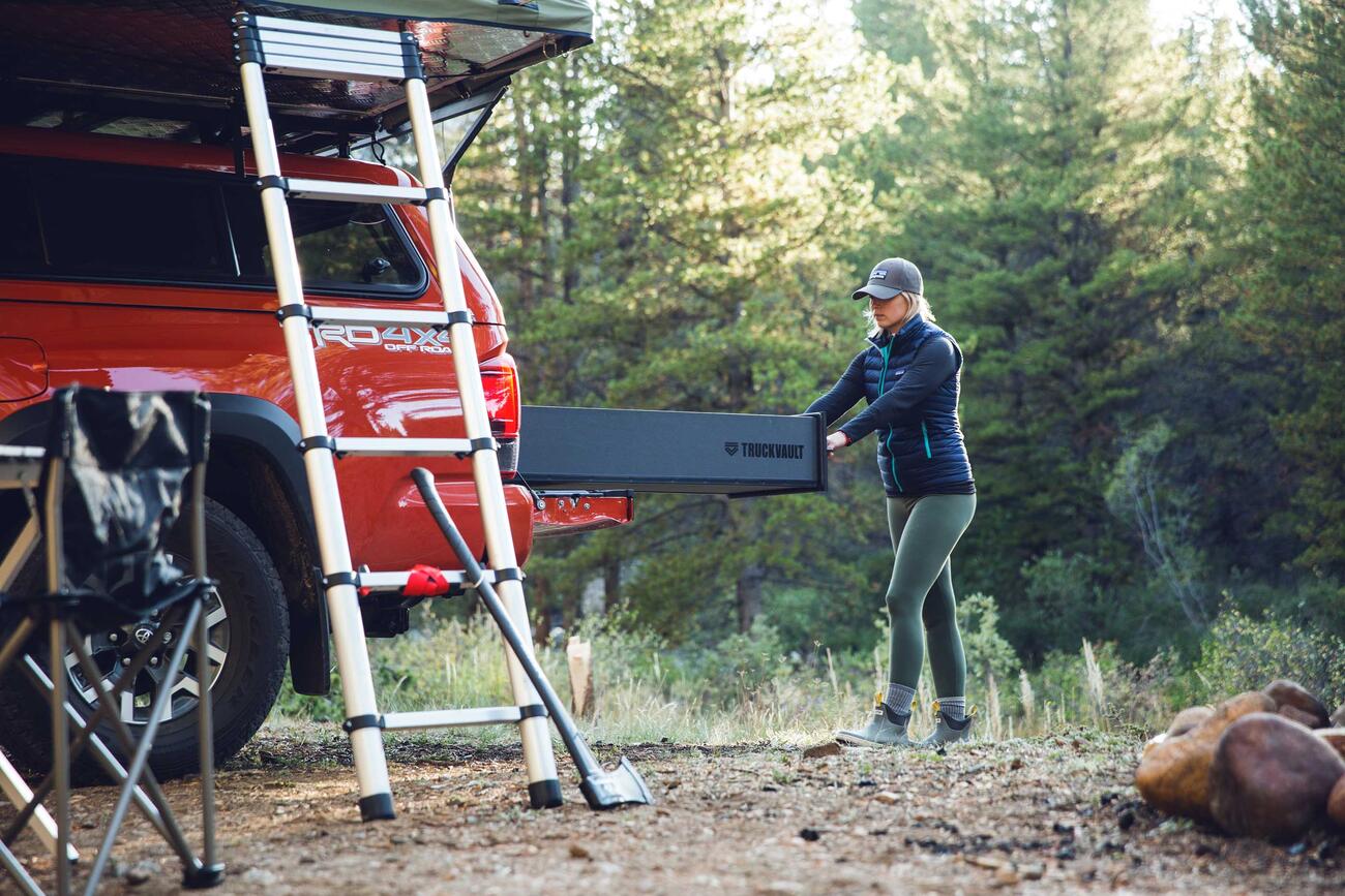 A woman opening a TruckVault in the back of a red truck, in the woods.