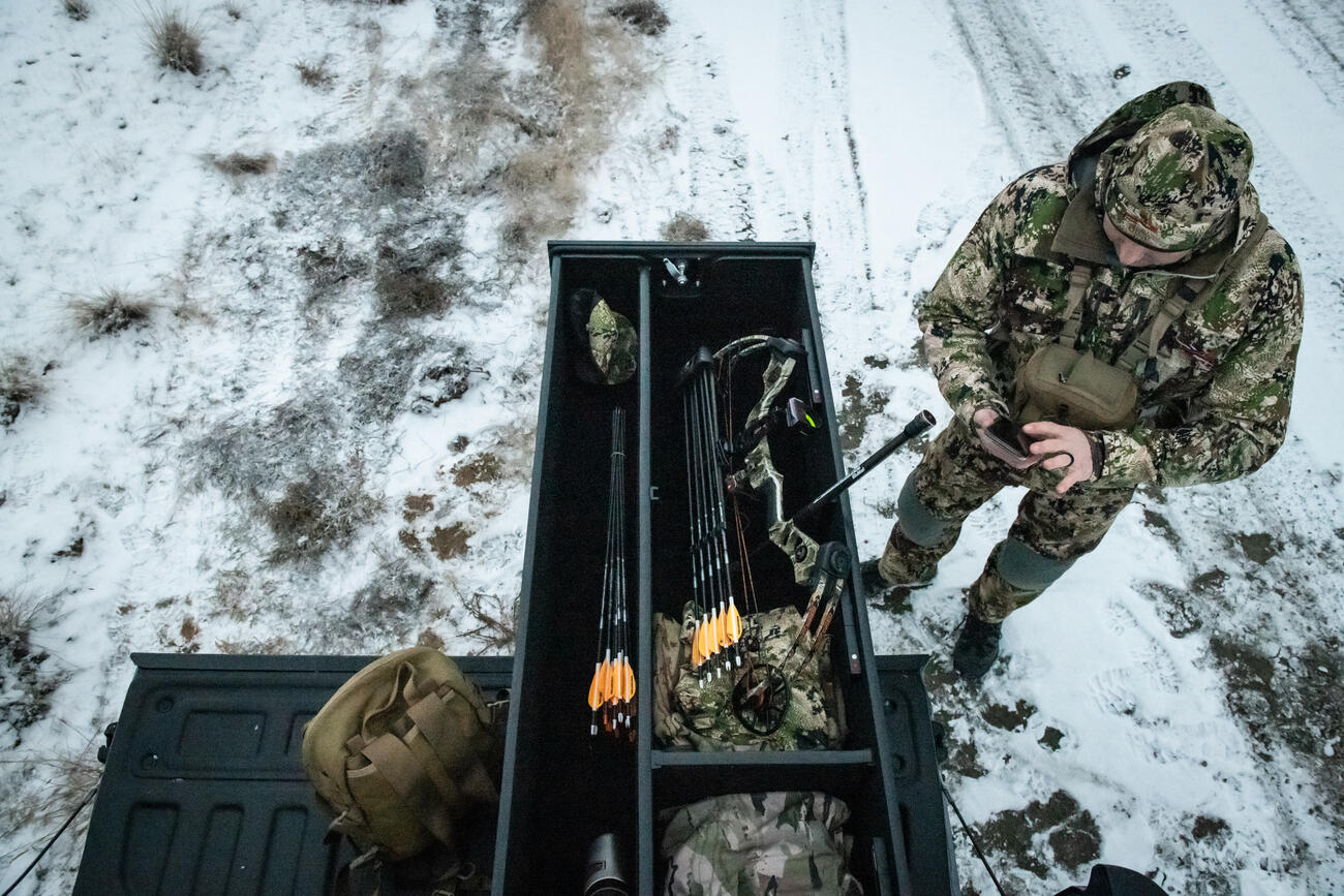 A man in camouflage checking his phone next to a TruckVault with a compound bow inside of it.