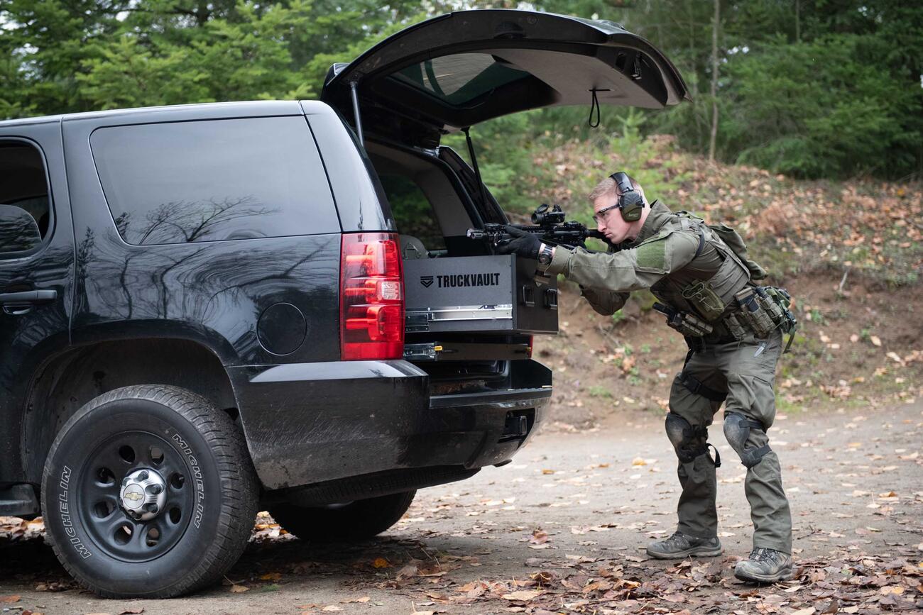 A man Shooting a gun that is resting on a TruckVault in the back of a black Chevy Tahoe.