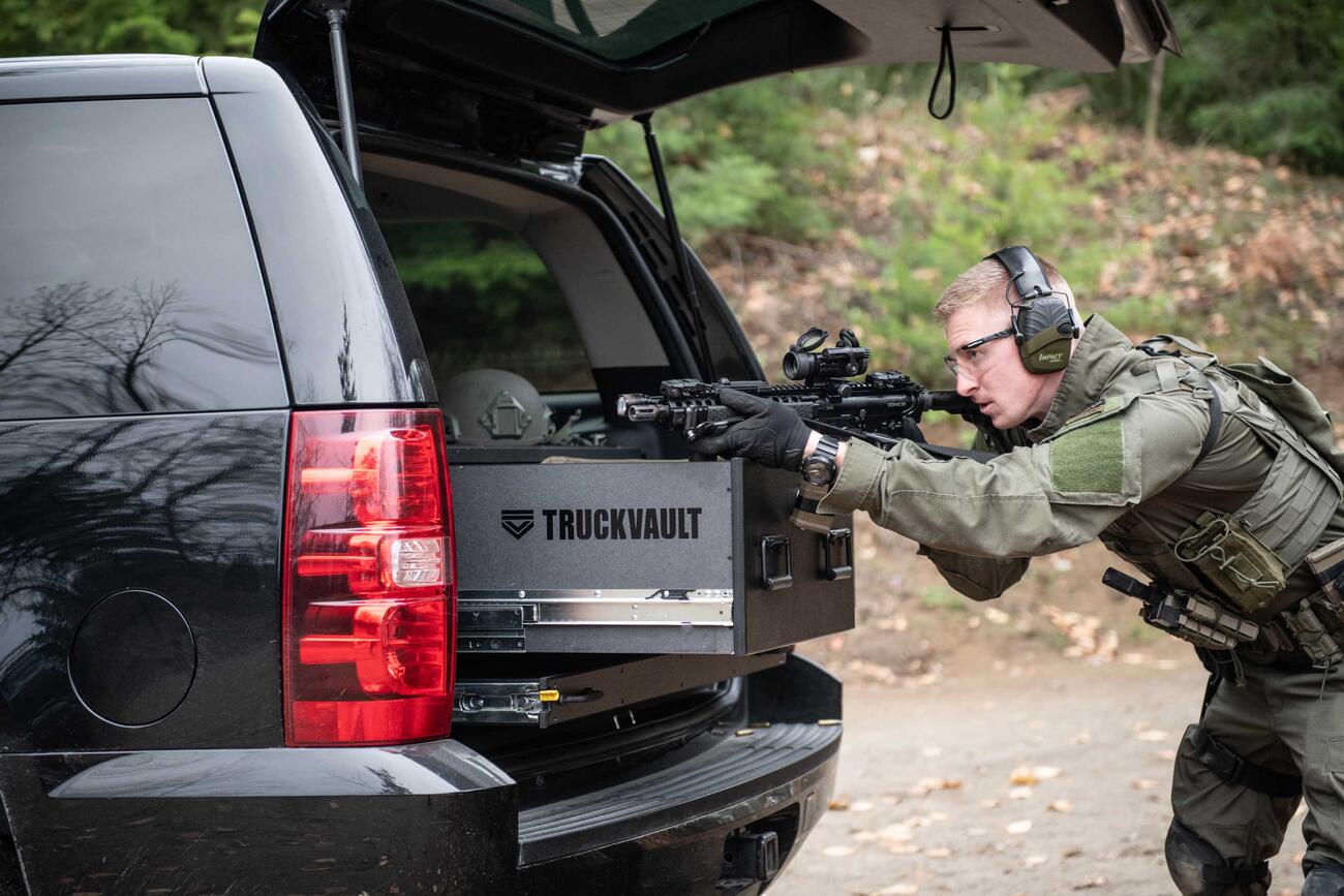 A man Shooting a gun that is resting on a TruckVault in the back of a black Chevy Tahoe.