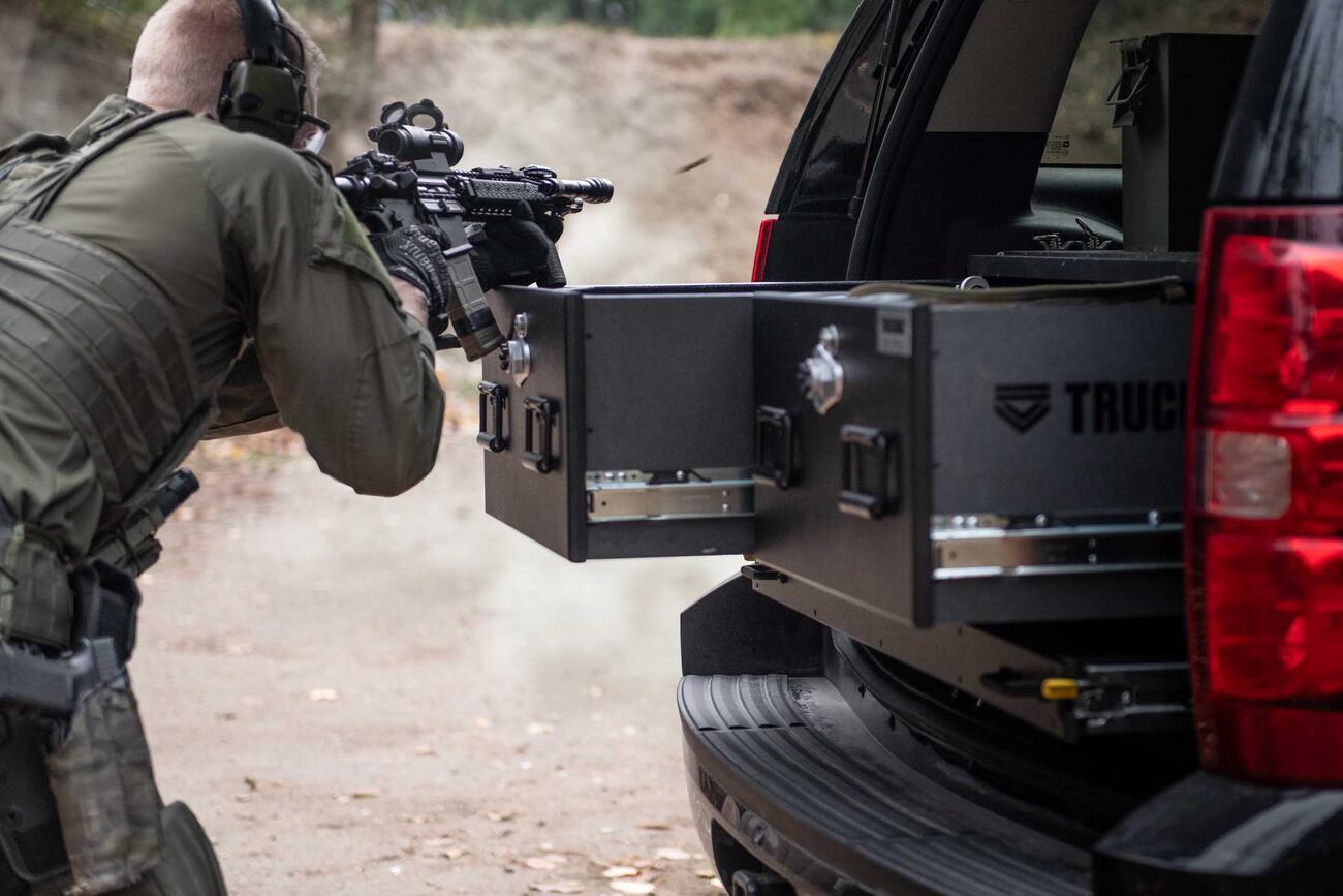 A man shooting a gun that is resting on a TruckVault in the back of a black Chevy Tahoe.