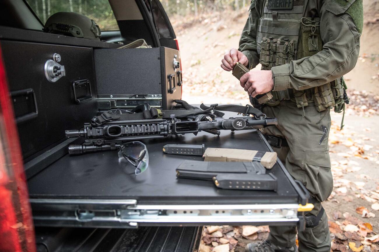 A man is loading his magazine while his rifle rests on a heavy duty pull out table in the back of a black Chevy Tahoe