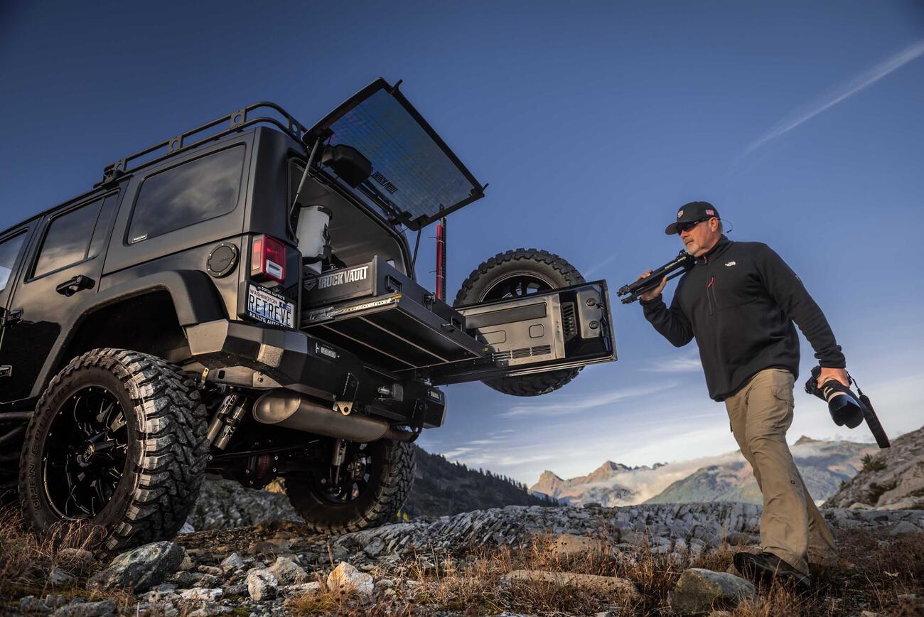 A man holding a camera and tripod near a black jeep with a TruckVualt in the mountains 