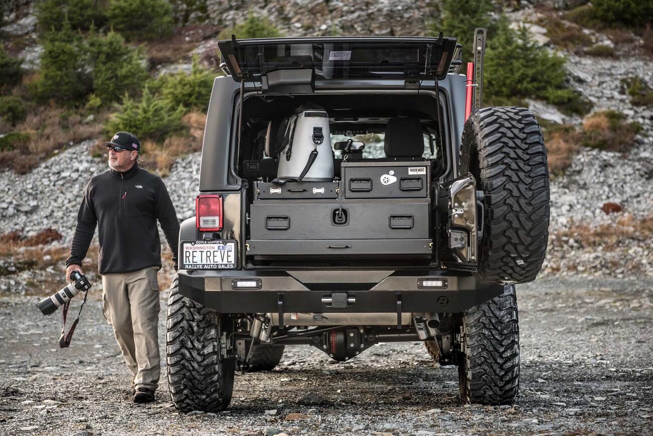 A man with a camera walking next to a 2013 Jeep Wranger with a TruckVault in the cargo space.