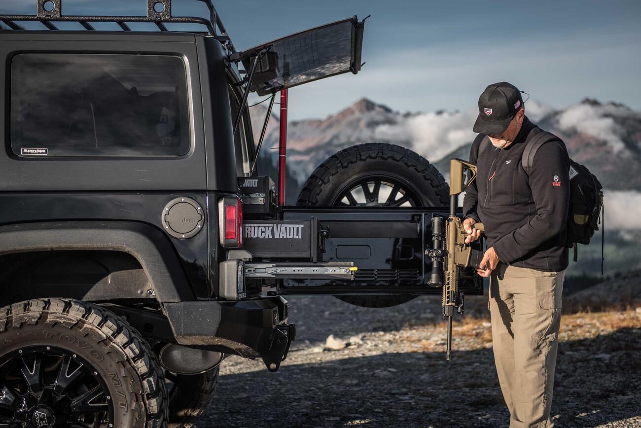 SUVseries_BaseLine_Custom_2013_Jeep_Wrangler_Unlimited_JK_DonFenton6.jpg A man holding an assault rifle behind a black Jeep Wrangler with custom TruckVault in the mountains.