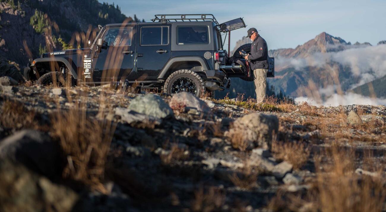 A man changing out a camera lense behind a black 2013 Jeep Wrangler with a TruckVault and a heavy duty pull out table in the mountains.