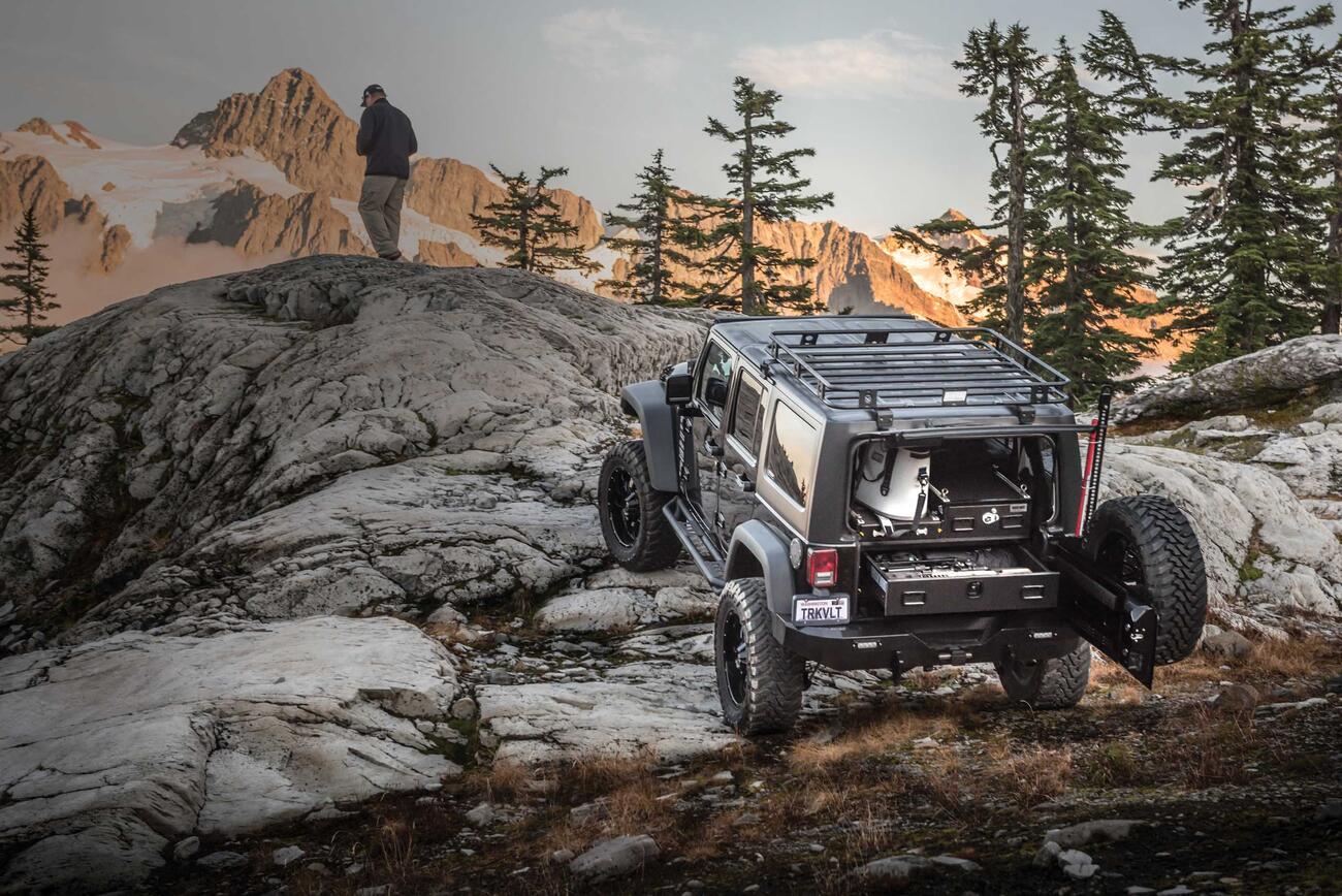 A gray Jeep Wrangler with a custom TruckVault parked on rocks in a mountain range with a man overlooking a valley.