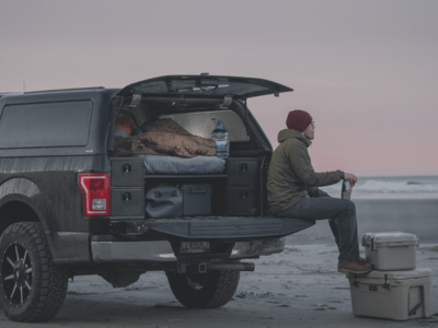 Black Ford F-150 with bed cap and tailgate open showing a TruckVault Base Camp 4. Man sitting on tailgate with feet resting on coolers watching the sun set at a beach
