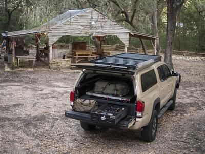 A Toyota Tacoma parked infront of a hut in the woods. Inside of the bed of the Tacoma is an open TruckVault filled with gear.