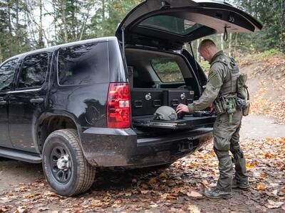 A man in tactical clothing working on a gun that is on top of a heavy duty pull out table in the back of a black Chevy Tahoe.