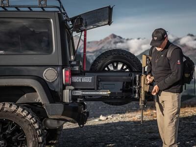 A man holding an assault rifle behind a black Jeep Wrangler with custom TruckVault in the mountains.